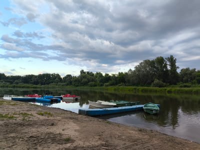 This media file depicts a serene waterscape at dusk in Ostrovnoye, Russia. The scene features a sandy embankment in the foreground, leading to a calm river. Several small boats are moored at a floating blue dock, including red pedal boats, white rowboats, and green motorboats. One green boat, numbered "16", is prominently visible. The river reflects the cloudy sky, which is a mix of light blue and darker grey, suggesting an overcast evening. A lush green forest lines the opposite bank of the river, adding to the natural beauty of the setting. The overall atmosphere is peaceful and tranquil, evoking a sense of calm as day transitions into night. There is no visible text other than the number on the boat.