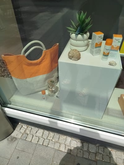 The image shows a display window of a shop in Offenbach am Main, Germany. The window contains a white display box with several items on it and in front of it. On the display box are two boxes of Eucerin Sun Protection Oil Control SPF 50, a bottle of Eucerin Sun Protection Oil Control SPF 50, a small green plant in a white pot, and two seashells. In front of the display box, to the left, is a large orange and white canvas tote bag with rope handles. To the right of the tote bag is a glass jar filled with coins. A small orange bottle, possibly a sunscreen sample, is placed on the floor in front of the tote bag. The display is set up on a white surface that is visible inside the window. The exterior of the display window shows a street scene with cobblestone pavement.