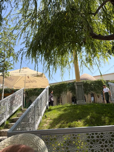 The image depicts a vibrant outdoor scene, likely a cafe or restaurant area, in Kuchtumgali, Uzbekistan, under a clear, sunny sky.
In the foreground, a large tree with abundant green, narrow leaves (possibly a willow) frames the left side. To the right, a white wicker chair is partially visible, indicating the viewpoint of the observer. A beige Coca-Cola branded parasol provides shade in the mid-ground.
Several decorative white metal screens with intricate geometric patterns, reminiscent of Islamic art, form a barrier or railing system, separating a grassy slope from what appears to be a paved or seating area. On the grassy incline, a person in a dark top and patterned skirt is visible. Further back, on a lower level near a building, another person in a white t-shirt and dark shorts stands.
The background features a building adorned with green ivy or vines, showing arched windows and brickwork. Behind this, a prominent domed structure with textured surface is visible against the blue sky, suggesting historical or traditional architecture. A tall wooden beam and strings of outdoor lights are also part of the setup.
The scene suggests a leisurely atmosphere, possibly an outdoor dining or relaxation spot, with natural elements complementing architectural details. The visible text is "Coca-Cola" on the parasol.