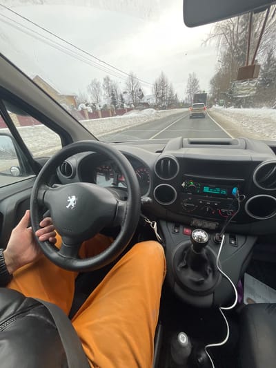The image presents a first-person, driver's perspective from inside a Peugeot vehicle, showing a winter road scene in Podvyaznovo, Russia. The driver's left hand, clad in a dark sleeve, rests on the black steering wheel, which features a silver Peugeot emblem. The driver's legs are visible, wearing orange trousers. The vehicle's interior includes a dark gray dashboard and center console with a manual gear shifter topped by a chrome knob. An infotainment system displays "05-23-21 Tue 8:37," and two blue USB chargers with attached white cables are plugged into the console. The windshield exhibits minor streaks and cracks, with a decorative object hanging from the rearview mirror.
Externally, a two-lane asphalt road is visible, partially covered with snow and slush on the shoulders and banks. Bare trees and residential structures with pitched roofs and fenced properties line the left side of the road. A light-colored SUV is observed traveling ahead in the same lane, with a darker, larger vehicle following further in the distance. The sky is overcast and grey. Overhead power lines are visible above the landscape.