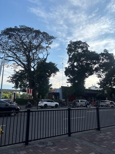 The image shows a street scene in Kelapa Dua, Indonesia. There are large trees lining the street, partially obscuring buildings. A KFC restaurant sign is visible behind the trees. Cars and motorcycles are parked along the street. A black metal fence runs along the side of the image. Above, the sky is blue with scattered white clouds.