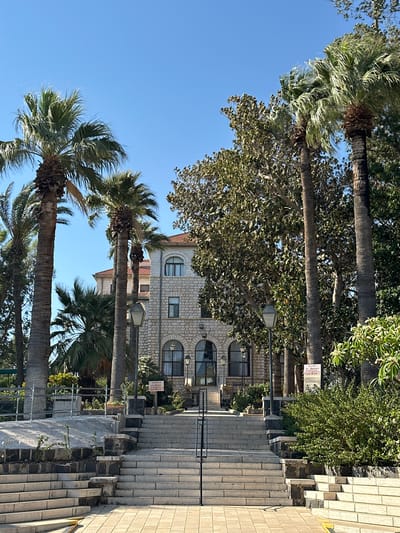 The image shows a wide stone staircase leading up to a two-story building with stone walls and arched windows. Palm trees and other foliage surround the building. A sign reading "ALTAR I ST. MATTHIEU" is visible near the top of the stairs. The scene appears to be a religious site in Emek HaYarden Regional Council, Israel, possibly the Mount of Beatitudes. There are no people or animals visible in the image. The sky is clear and blue, suggesting a sunny day.