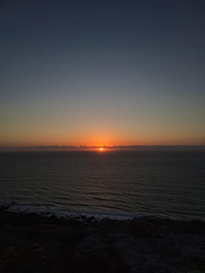 A vertical shot captures a serene sunset over the ocean, likely near Ahrara, Morocco. The sky transitions from a deep navy blue at the top to a warm orange glow near the horizon. A thin band of clouds stretches across the ocean, partially obscuring the setting sun. The sun itself is a bright orange orb, just touching the cloud line. The ocean below is calm, with subtle ripples reflecting the fading light. In the foreground, a dark, rocky coastline is visible, with waves gently breaking against the shore. The overall scene is tranquil and evokes a sense of peace as day turns to night.