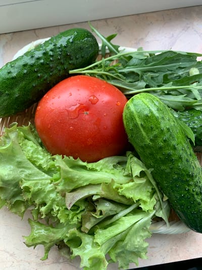 A top-down, close-up shot features a wicker basket filled with fresh vegetables and greens. The basket is partially visible at the bottom and right edges of the frame. Occupying the majority of the basket are two bumpy, dark green cucumbers, one positioned horizontally across the top and another vertically on the left side. Both cucumbers appear to have water droplets on their skin.
In the center of the basket is a large, bright red tomato, also glistening with water droplets. To the right of the tomato and below the top cucumber, a cluster of vibrant green lettuce leaves is prominently displayed. The lettuce has a ruffled texture. Behind the tomato and to the left, a bunch of dark green arugula leaves, with their characteristic lobed shape, are visible.
The background consists of a beige surface with a subtle marbled pattern. The lighting suggests an indoor setting, possibly near a window, given the soft illumination and the presence of what appears to be a windowsill or countertop. There is no visible text or indication of specific time of day or weather. The overall impression is of a fresh harvest or ingredients prepared for a meal.