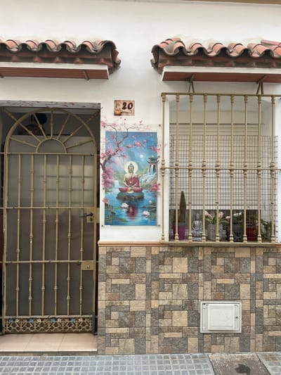 The image shows the facade of a building at number 20 in La Línea de la Concepción, Spain. A metal security gate is visible on the left, with a decorative pattern at the bottom. To the right of the gate, a large painting of Buddha in meditation, seated on a lotus flower in a body of water with cherry blossoms and a pagoda in the background, is displayed on the white wall. A window with a metal security grille is situated to the right of the painting. Inside the window, several potted plants, including a cactus and decorative Buddha statues, are visible. The lower part of the building is covered with a stone tile pattern, and a white electrical or utility box is mounted on the wall. The roof is tiled with traditional red Spanish tiles. The street in front is paved with grey cobblestones.