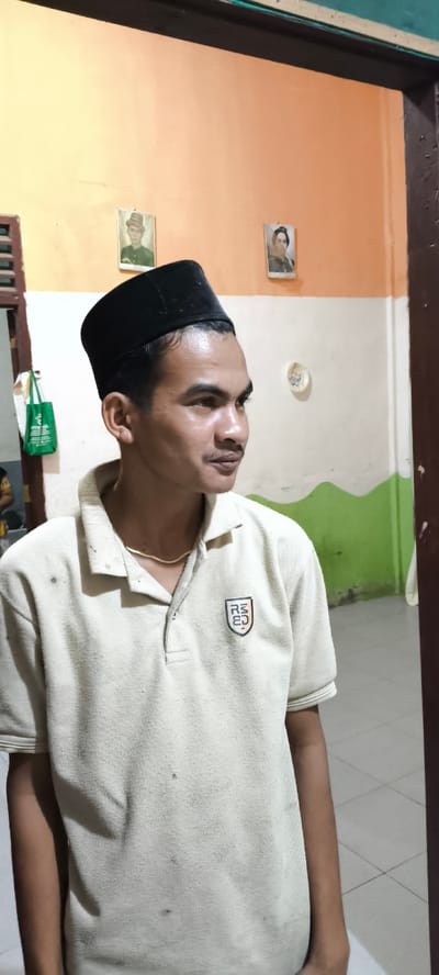 The image depicts a medium shot of a man wearing a black peci (a cap) and a light-colored polo shirt with a small crest on the left chest. He is looking to his right, with a slight smile. The background suggests an indoor setting, possibly a home, with a wall painted in orange and white with a green wavy pattern. Two framed pictures are visible on the orange section of the wall. To the left of the man, a green tote bag hangs on a door frame. The floor appears to be tiled. The image quality is fair, with some graininess. The scene is likely situated in Aluebutuha, Indonesia, based on the context provided.