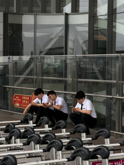 Three young men in white shirts and black ties are sitting on the ground in front of a modern building with large glass windows. They are all looking at their mobile phones. In front of them are rows of airport luggage carts. The scene appears to be outdoors, possibly at an airport or train station, given the presence of the carts and the modern architecture. The lighting suggests it is daytime. The building's reflective windows show a blurred reflection of the sky and possibly other structures. There is a railing in front of the men, and further in the background, more reflections are visible in the glass. One of the men has glasses and is smoking a cigarette. There is signage on the glass panels behind the men, some of which appears to be in Chinese. The overall atmosphere is one of waiting or downtime.