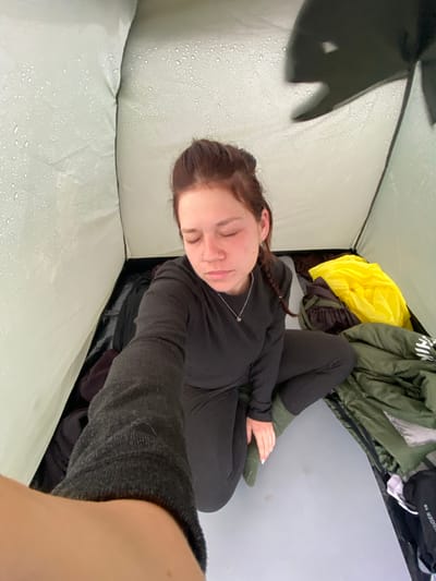 A high-angle, medium shot shows a young woman sitting inside a tent, with her eyes closed and her head tilted down. She has auburn hair pulled back in a braid and is wearing a black long-sleeved shirt and black pants, with green socks. The tent's interior is visible, with water droplets on the fabric. To her right are camping supplies, including a green sleeping bag and a bright yellow bag. The floor of the tent is a light grey material. The scene is set in the natural landscape of Абзаново, Russia, which is evident from the context of camping.