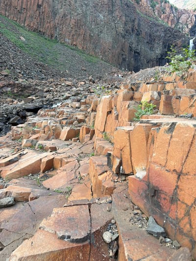The image shows a rocky, natural landscape with evidence of human activity. In the foreground, large, angular orange rocks are stacked and broken, with some small green plants growing in the crevices. Further back, a steep slope of smaller grey rocks is covered in sparse green grass and some purple wildflowers. Behind this, a towering cliff face is composed of dark, vertical rock formations, resembling basalt columns. In the distance, a waterfall can be seen cascading down the cliff. Several small groups of people are visible on the slope and near the cliff face. On one of the cliff faces, there is a marking of "6418" in white paint. The scene suggests a location accessible to hikers and visitors, likely a natural park or reserve. This geological formation is characteristic of the Talnakh ore-bearing region near Norilsk, Russia, known for its unique columnar basalt formations.