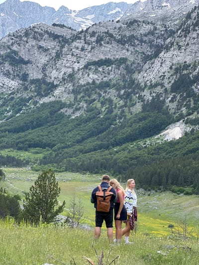 Three individuals are standing in a grassy area with a mountainous landscape in the background. The person on the left is a male wearing a dark blue jacket with a brown backpack and black shorts. The person in the middle is a female wearing a pink top and a black skirt. The person on the right is a female wearing a white and gray patterned cardigan over a white top, a red and black plaid skirt, and white socks. They are facing away from the camera, looking towards the valley. The environment is characterized by lush green grass, scattered trees, and a vast, rocky mountain range with patches of snow at the higher elevations. The lighting suggests it is daytime, likely late morning or early afternoon. The location is identified as being in Shalë, Albania, characterized by its rugged mountain terrain and natural beauty.