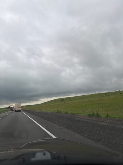 The image depicts a view from inside a vehicle driving on a highway, likely in the vicinity of Nizhniye Bishindy, Russia, given the context. The sky is overcast with heavy, grey clouds, suggesting impending or ongoing inclement weather. The highway is asphalt, with a white lane divider visible. To the right of the highway, a grassy hillside slopes upwards, dotted with sparse vegetation. Two trucks are traveling ahead on the highway. The vehicle's dashboard is partially visible in the foreground, reflecting the cloudy sky. The overall scene is one of a journey through a rural, possibly isolated, landscape under a somber sky.