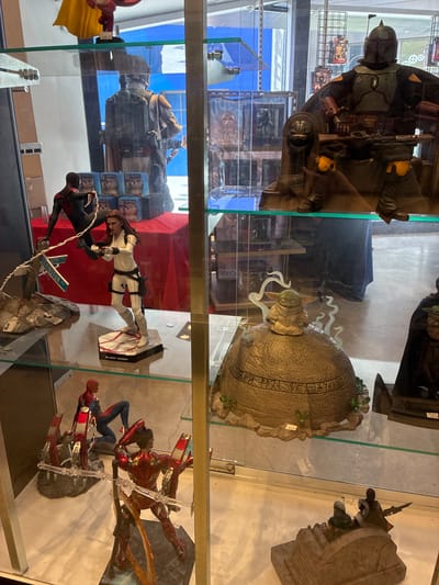 A medium shot shows a glass display case filled with collectible action figures and statues. The figures are arranged on multiple shelves and pedestals. The top shelf features a large figure of Boba Fett seated on a throne. Below that, on the second shelf, a Black Widow action figure is displayed in a dynamic pose, holding a gun, with a Spider-Man figure in the background. The third shelf showcases a Grogu (Baby Yoda) figure perched on a decorative, weathered dome with an inscription. Further down, on the bottom shelf, a scene with Iron Man and Spider-Man figures is visible, and another group of figures, possibly from The Mandalorian, are on a vehicle. The background is blurred, but glimpses of shelves, posters, and lighting suggest an indoor retail environment, possibly a comic book or collectible store. The overall lighting is bright, illuminating the glass case and its contents. The location is identified as Unknown, United States.