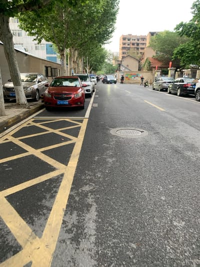 The image shows a street scene in Wuhan, China, located at latitude 30.508, longitude 114.404. There is a line of parked cars along the right side of the road, with a red car at the forefront and a silver car behind it. The road is empty with a man riding a motorcycle in the distance. There is a man walking on the sidewalk. There is a drain cover in the middle of the road. The street is flanked by trees on either side, with a multi-story building on the left side and a smaller building on the right. The pavement is asphalt and there are yellow markings on the road. The weather appears to be overcast and there is a light rain falling.