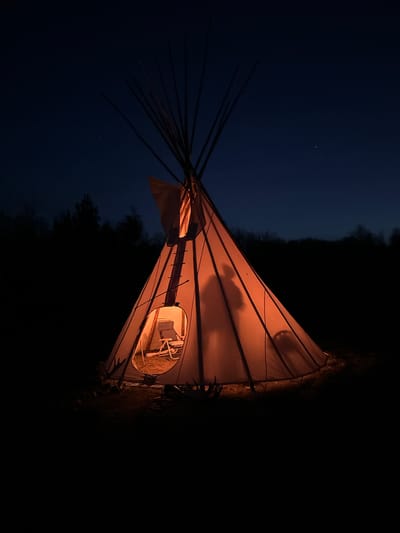 A low-angle shot depicts a teepee at night. The interior of the teepee is lit, with a chair visible through the doorway. Dark trees are visible in the background and a very dark blue sky. The location is specified as Unknown, Canada.