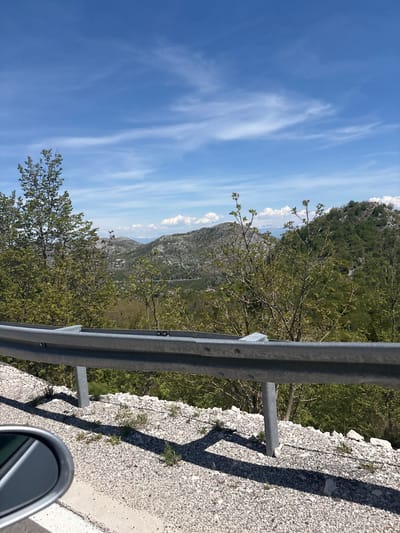 The image, taken from a vehicle, depicts a sunny mountain landscape. A metal guardrail runs horizontally across the mid-foreground, separating the viewer from a steep, tree-covered slope. The perspective suggests the vehicle is on a winding road or viewpoint high up.

In the immediate foreground, to the right of the guardrail, is a gravel-like road shoulder with scattered small plants. Long, distinct shadows are cast across this rocky surface, indicating bright sunlight, likely mid-day or early afternoon. A portion of a car's side mirror is visible in the bottom right corner, reinforcing the perspective from inside a moving vehicle.

Beyond the guardrail and the immediate slope, lush green trees with fresh spring foliage dominate the mid-ground. Further in the background, rugged, greyish mountains stretch across the horizon, partially covered in vegetation. Some white, fluffy clouds are visible along the mountain peaks.

The sky is a clear, vibrant blue, occupying the upper portion of the image. Wispy, white cirrus clouds streak across the expanse, adding texture to the bright sky.

The scene conveys a sense of travel through a scenic, mountainous region. Given the provided context, the location is Obzovica, Montenegro. No visible text is present in the image.