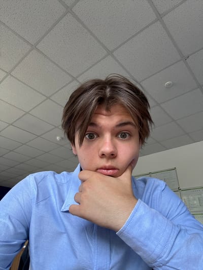 The image depicts a young male individual, appearing to be Caucasian, with medium-length brown hair, captured in an indoor setting. He is looking directly at the viewer with wide, attentive eyes. His left hand is positioned under his chin, with knuckles resting against his lower lip, suggesting a pensive or observant posture. He is wearing a light blue, long-sleeved button-up shirt. The background features a white suspended acoustical tile ceiling with a visible grid pattern and a circular smoke detector or light fixture in the upper right. To the subject's right, a light-colored wall is visible, with several sheets of paper or charts, possibly with green borders, affixed to it, indicating an office or classroom environment. The image does not contain specific geographical markers to confirm its precise location within Mamhilad, United Kingdom.