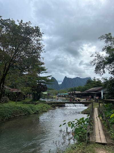 The scene depicts a riverine recreational area in Ban Nampoun, Laos, under a heavily overcast sky with dense grey cloud cover. In the background, prominent karst limestone mountains rise, their peaks partially obscured by atmospheric haze or clouds.
A clear river flows from the background towards the foreground, featuring a man-made weir that creates a small waterfall in the mid-ground. Dense green foliage and large trees line both riverbanks.
On the left bank, multiple rudimentary wooden huts with tin roofs are visible amidst thick vegetation. Several individuals, some appearing to be in swimwear, stand near these structures and at the water's edge, interacting.
The right bank features a longer, dark-roofed wooden building and a series of smaller, uniformly constructed wooden bungalows with pitched roofs, some appearing to be changing rooms or seating areas. Numerous individuals are engaged in water activities, swimming or wading in designated pools or sections of the river. Others are congregated on wooden platforms that extend over the water, socializing. A simple wooden plank bridge with timber posts spans the extreme right foreground, connecting to an area of overgrown vegetation. Small boats or rafts are visible further upstream in the river.