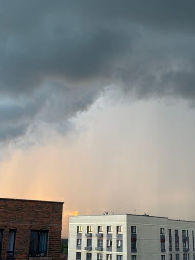 The image depicts a sky with dramatic, dark clouds dominating the left side and a bright, hazy sky on the right, suggesting either sunset or sunrise. Two modern apartment buildings are visible on the right. The taller building on the far right has a light-colored facade, possibly white or light gray brick, with numerous windows. The building in the foreground on the right is clad in red brick. The sky is a mix of dark grays and lighter tones, with a warm, golden hue near the horizon on the right, potentially indicating the sun peeking through or a reflection. There is no discernible activity of people, and no visible text. The scene is set in Laikovo, Russia. The overall impression is one of atmospheric change, with a storm potentially approaching or receding, juxtaposed against urban architecture.