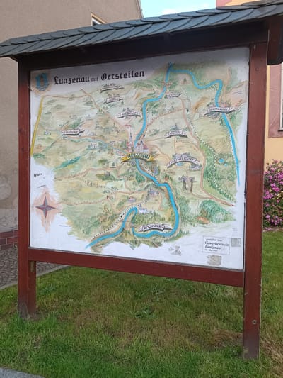 The image shows a framed map titled "Lunzenau mit Ortsteilen" (Lunzenau with Districts) standing on grass in front of a building. The map depicts the town of Lunzenau and its surrounding areas in Germany. It includes various locations such as HIMMELHARTHA, ELSDORF, SCHLAISDORF, GROSS-SCHLAISDORF, GOSSEN, HOHEN-KIRCHEN, BERTHELSDORF, ROCHSBURG, and GORITZHAIN. The map features a compass rose and an inscription stating it was donated by the Gewerbeverein Lunzenau in May 1999. A portion of the map’s artist signature, D. Steinbach 1999, can be seen. The frame is made of wood with a dark red finish. The surrounding environment includes a building with beige and red walls, greenery, and blooming pink flowers.