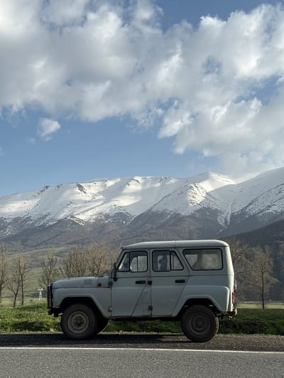 A white UAZ-452 van sits parked on the shoulder of a gray asphalt road in Margahovit, Armenia. The road runs parallel to a green grassy area interspersed with bare trees. Behind the van, a large mountain range, partially covered in snow, dominates the landscape, with a clear blue sky and puffy white clouds above the mountain.