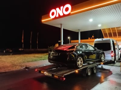 At night, a black Tesla Model S is loaded onto a car carrier trailer. The trailer is parked under the canopy of a gas station with a prominent "ONO" sign illuminated in red. A white van is parked to the right of the trailer. The ground is wet, reflecting the lights. In the background, several other vehicles are visible, and the area appears to be a roadside service station or rest stop. The license plate on the trailer reads "BT 08 EXE". The location is indicated as Blatnice, Czechia.