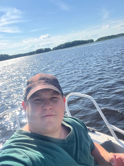 The image shows a person taking a selfie while on a boat. The person is wearing a camouflage baseball cap and a green t-shirt. They have a slight smile on their face. The boat appears to be on a body of water, possibly a lake or river, with trees lining the shore in the background. The water is rippling and reflecting the sunlight, creating a sparkling effect. The sky is blue with some scattered clouds. The sun appears to be high in the sky, suggesting it is daytime, likely late morning or early afternoon. The overall mood seems relaxed and enjoyable, indicating the person is having a pleasant time on the water. There is no visible text in the image. The location context provided is Сюктерка, Russia.