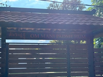 The image shows a gated entrance. The gate is constructed of horizontal wooden slats set within a dark metal frame. Above the gate, a dark wooden board displays text in two languages or scripts. On the left, is Tibetan script, while on the right, the phrase "OM MANI PADME HUM" is written in English. Above the sign, is a red and brown tiled roof. Trees are visible through the gate and above the roof. The location is presumably in Ciudad de la Costa, Uruguay, as per the prompt's instruction.