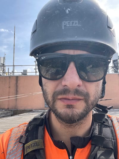A man wearing safety gear, including a Petzl-branded helmet and harness, sunglasses, and an orange safety vest, poses for a selfie outdoors. Behind him are various telecommunications infrastructure and a light-colored building under a blue sky with some clouds. It is not possible to confirm the location as Hertsmere, UK, from the image provided.