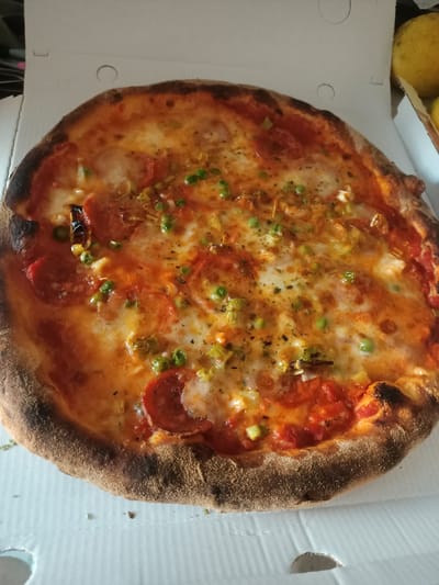 The image shows a close-up, overhead view of a whole pepperoni pizza inside a white cardboard pizza box. The pizza has a thick, slightly charred crust, and is topped with melted cheese, tomato sauce, pepperoni slices, green peas, and what appears to be chopped leeks or scallions. The pizza box is open and rests on a surface that appears to be a table. In the upper right corner of the image, partially visible, are two yellow fruits, possibly lemons or oranges. The background is blurred, but suggests an indoor setting. There are no people visible in the image, and no specific landmarks or clues within the image itself that would indicate the city of Riposto, Italy, or any other specific location.