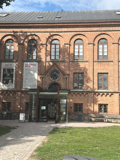 The image shows the exterior of the Historical Museum (Historiska Museet) in Lund, Sweden, a red brick building with arched windows and a dark metal roof. Two banners are hung on the facade. The banner on the left is titled "SKATTER FRÅN FEM KONTINENTER" (Treasures from Five Continents) and displays images of artifacts. The banner on the right, "HISTORISKA MUSEET," welcomes visitors to the museum and provides opening hours. The entrance to the museum is visible, featuring glass doors with a decorative motif. In front of the museum, there is a paved walkway leading to the entrance, flanked by grass and trees. Sunlight casts shadows on the building and the ground.