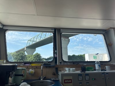This media file captures a view from inside a boat's cabin, looking out through two large windows. The scene is set during the daytime, under a bright, cloudy sky. Lush green trees line the banks of a waterway, and a massive concrete bridge, likely a highway or railway bridge, spans across the view. The bridge's structure is prominent, with large concrete pillars supporting the elevated roadway. In the right window, a large, white object, possibly a life raft or storage container, is visible on the bank.

Inside the cabin, a functional area is visible. On the left, a coffee machine sits on a counter next to a green electric kettle. A sink with a silver faucet is also present. Further to the right, a collection of mugs, a box of tea, and a bottle of water are arranged on a control panel. The panel has various dials and buttons, indicating this is a control area of the boat. Two white cups are neatly placed on the counter, suggesting a moment of rest or a break.

The overall atmosphere appears calm and perhaps a bit utilitarian, as is common in boat cabins. The time of day is clearly daytime, indicated by the bright sunlight and the clear visibility. The weather appears fair, with scattered clouds in a blue sky. The presence of the large bridge and the waterway strongly suggests a location near a significant river or canal, consistent with Brunsbüttel, Germany, a port town known for its canals and bridges. No people are visible in the image.

Visible text includes "SOLARGLIDE" and "MS" on the window blinds. On the bottle of water, "AQUA" and "STILL" are legible. A small label on the control panel shows a symbol of a lightning bolt in a red triangle. A mug with a yellow sun and text "you are my sunshine" is also visible.