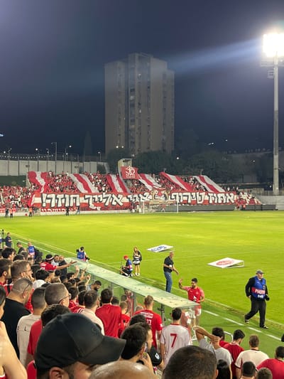 The image captures a nighttime scene inside a sports stadium, identified as the Green Stadium (Tuvia Bahr Stadium) in Nof HaGalil, Israel. A significant crowd of spectators is visible, primarily concentrated in the foreground and a section of the main stands.

In the foreground, numerous individuals, predominantly male, stand behind a clear barrier, their backs or profiles towards the viewer, observing the field. Many wear red or white shirts. On the immediate edge of the bright green grass field, a man in a red jersey faces the crowd at the barrier, with his right hand raised. Nearby, a man in a black shirt and blue jeans stands, and a woman in a black and white striped top is crouched. Further along the field perimeter to the right, a person wearing a blue vest with "PHOTO 048" printed on the back walks from right to left. Two white, angled advertising boards featuring red logos rest on the grass.

The main stadium stands are densely populated with fans, most wearing red or white, displaying large, horizontally striped red and white banners. A prominent white banner with black Hebrew text, "אתם אלינו, אנחנו חזק!" (You to us, we are strong!), is stretched across the lower part of these stands. In the background, beyond the stadium structure, a tall, multi-story concrete building with visible windows rises against the dark blue-black night sky, partially obscured by trees. A bright stadium floodlight tower on the far right illuminates the scene, casting a strong light beam upwards.
