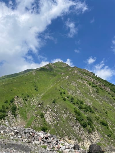 The image shows a vast, green mountain slope under a partly cloudy blue sky. The mountain is covered in lush grass, with scattered trees dotting its surface. Rocky outcrops and ravines are visible on the mountainside, creating a textured landscape. In the foreground, a pile of rocks and debris is present, suggesting a rugged, natural environment. The scene appears to be in a mountainous region of Almasiani, Georgia, characterized by its dramatic natural scenery. There are no people or animals visible in the image.