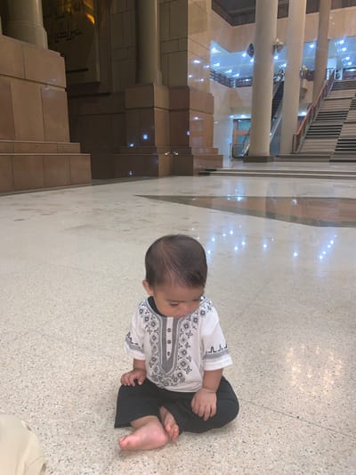 The image, rotated clockwise, shows a baby sitting on a light-colored, speckled floor in what appears to be a large, modern indoor public space, possibly a mosque or a commercial building in Samarinda, Indonesia.

The baby is the central subject, dressed in a white long-sleeved shirt featuring a black and grey geometric pattern on the chest, collar, and cuffs, paired with black pants. The baby is sitting with legs bent, facing slightly to the right, looking down or forward. Bare feet are visible.

The background reveals a multi-level structure characterized by tall, smooth, light-colored pillars. Beyond the pillars, various architectural elements are visible, including several flights of stairs and an escalator, indicating a multi-story building. The floor and some wall surfaces are highly polished, reflecting numerous bright points of artificial light from overhead fixtures. Some sections of the building's interior walls are clad in darker, possibly brown, materials, creating visual contrast. On a distant dark wall, there appears to be some indistinct, light-colored writing or calligraphy, but it is too blurry to decipher. The overall lighting is bright and artificial, suggesting an indoor setting at any time of day. The baby appears calm, engaged with something outside the frame or looking downwards.