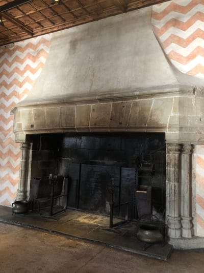 This image shows a large fireplace in the Chillon Castle, located in Veytaux, Switzerland. The fireplace is constructed of stone and blackened from soot, with visible stone detailing above the firebox. Inside the firebox are metal racks and what appear to be cooking pots on stands. The surrounding walls feature a salmon and white chevron pattern above and on the left side.