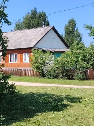 The image shows a rural scene with a wooden house, a fence, and trees under a clear blue sky. The house is light brown with white window frames and a grey corrugated roof. A wooden fence runs along the front of the property, enclosing a grassy yard. Several trees, including some with green foliage and others with bare branches, are visible behind the house and fence. The ground in the foreground is covered with short green grass. The image appears to be taken on a sunny day.