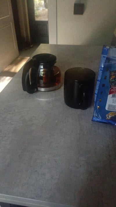 The image shows a countertop in what appears to be a domestic setting. On the countertop, there is a glass coffee pot with a black handle, filled with a dark liquid, possibly coffee or tea. Next to the coffee pot is a black mug. To the right of the mug, a portion of a blue package is visible, with some text and graphics on it, including what appears to be a cartoon character. The countertop itself is speckled grey. In the background, a doorway or window is visible, leading to an outdoor area with trees. A white door is partially visible on the left side of the image. The lighting suggests it is daytime. There are no people visible in the image. The location of Nikolino, Russia is not explicitly depicted or inferable from the visual information in the image.