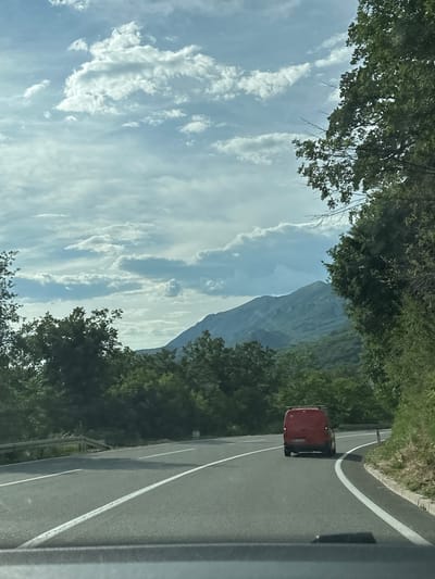 Captured from a moving vehicle on a road near Golubovići, Montenegro, the image depicts a landscape dominated by a mountain range under a partly cloudy sky. The road, marked with white lines, curves to the right. A red van travels on the road. Trees line both sides of the road, partially framing the mountain view.