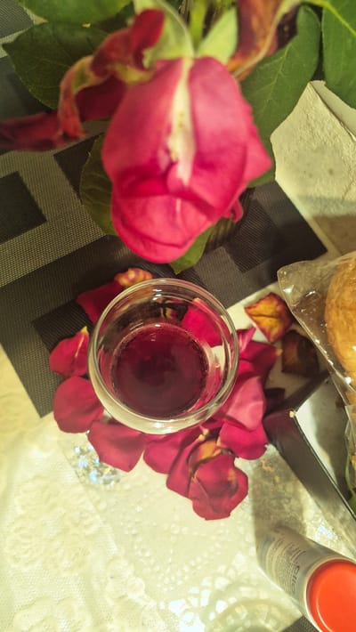 This is a top-down, close-up, still image of a table setting. The main subjects are a partially opened pink rose, scattered red rose petals, and a small glass filled with a dark red liquid, likely wine. The rose is in the foreground, to the left, with its green leaves visible. The glass of wine is in the center, surrounded by rose petals.

In the background, there is a dark rectangular box, possibly a food container, and part of a round, golden-brown item, perhaps a pastry or bread, enclosed in clear plastic. To the right of the box is a cylindrical container with a red cap. Visible text on this container appears to be "ello" and further text in a smaller font.

The objects are arranged on a dark patterned placemat, which is partially covered by a light-colored, possibly lace, tablecloth. The lighting suggests an indoor setting, and there are no clear indicators of time of day or weather. The overall mood is romantic or celebratory.

The setting is Ialoveni, Moldova.