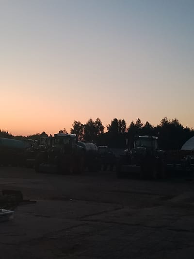 The image captures a serene sunset scene in Nova Basan, Ukraine, with agricultural equipment silhouetted against a gradient sky transitioning from pale blue to soft orange. The foreground is dominated by several tractors and what appear to be agricultural implements, possibly parked after a day's work. The lighting suggests it is either sunrise or sunset, casting long shadows and creating a somewhat moody atmosphere. The trees in the background form a dark, dense line against the horizon, with some lighter foliage visible. To the right, a portion of a large, curved structure, possibly a storage building or greenhouse, is visible. The ground appears to be a dirt or gravel lot, with some scattered debris and subtle textures. There are no people visible in the frame, and the overall impression is one of quietude and the end of a working day in a rural setting. The time of day is clearly indicated by the twilight sky.