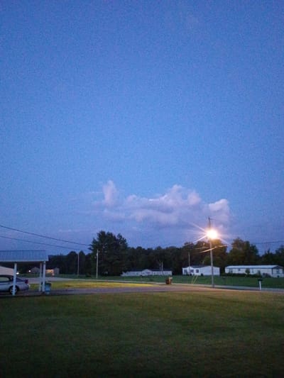 The image shows a twilight scene in a suburban area of Jeffersonville, United States of America. The sky is a deep blue, with some scattered clouds. A street light is illuminated, casting a bright glow. In the foreground, there is a grassy lawn. To the left, a silver car is parked under a carport. In the background, there are several mobile homes and trees. The overall impression is one of a quiet, peaceful evening.