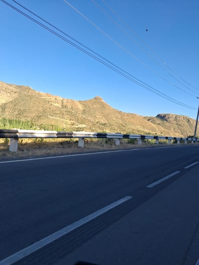 The image displays a road with a guardrail and a mountainous landscape under a clear blue sky. The road is dark asphalt with white lines marking the lanes. The guardrail is metallic with alternating black and white reflective stripes, supported by concrete posts. Beyond the guardrail, there is dry, sparse vegetation and then the sloping hills of the mountain range. The mountains are arid, with patches of green vegetation on their lower slopes, and appear to be made of rock and earth. In the sky, there are power lines stretching across the frame, and a small black bird is visible in flight high above. The scene suggests a rural or semi-rural environment. The prompt mentions the city of Shatin, Armenia, but there are no specific visual cues in the image to confirm or deny this location.