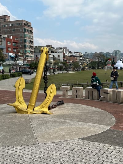 A gold anchor stands prominently on a concrete structure, with a chain visible. Behind it lies a grassy area leading to buildings. People are seen, including some interacting with large, blocky lettering that spells out "TAMSU." The scene is partially sunny with scattered clouds. The anchor is on the right side of the image. The concrete is gray and the brick pavers are a mix of red and gray.