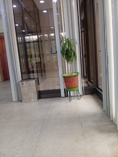 A metal trash receptacle, a potted plant on a wrought iron stand, and vertical blinds are situated on a terrazzo floor, adjacent to a glass door. The scene is likely located in Shiroro, Nigeria.