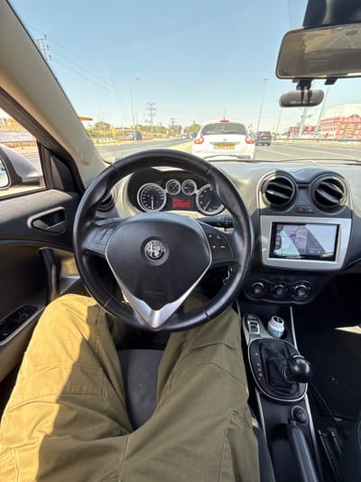 The image shows the interior of a car. The driver, wearing green cargo pants, is driving with their right hand on the steering wheel. The car is an Alfa Romeo, with the steering wheel showing the Alfa Romeo logo. The dashboard shows the speedometer and tachometer, as well as a navigation system. There is a white bottle in the center console. The car is in motion, with the view of the road outside the windshield showing other vehicles and buildings. The location is on a road in Kiryat Ekron, Israel, at coordinates 31.865, 34.814.