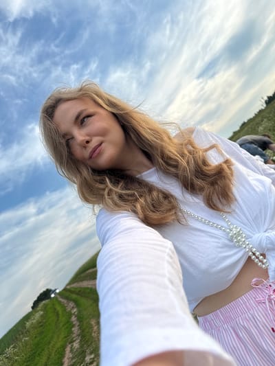 A young woman with long blonde hair is taking a selfie outdoors. She is wearing a white knotted top, pink striped pants, and a pearl necklace. The background shows a field with a path leading up a hill, a car in the distance, and a cloudy blue sky. Another person is visible in the background lying in the field. The location appears to be a rural area.