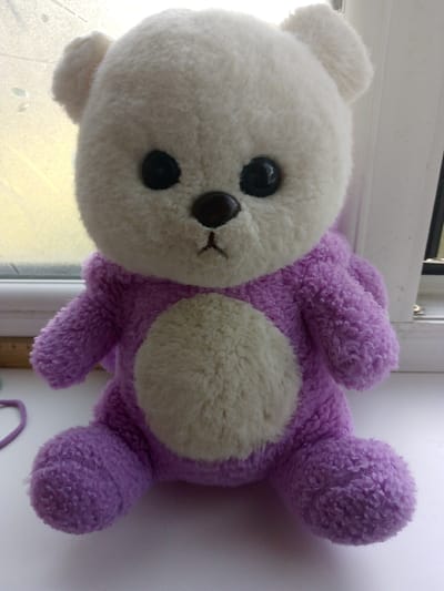 A white and lavender plush teddy bear is sitting on a windowsill. The bear has a white head and a lavender body. Its eyes are black, and its nose is brown. The bear is sitting in front of a window with a yellow-tinted light filtering through. To the left of the bear, a small section of a purple string is visible. The windowsill is white, and a ruler with dark markings is partially visible at the bottom left of the frame, resting on the windowsill. The background to the right of the bear shows a white window frame and a dark vertical element, possibly a window pane. The setting is indoors, with natural light coming from the window. The overall scene appears to be a domestic setting. It is not possible to determine the location in the city of Prigorognoye, Kyrgyzstan, from the visual information provided.