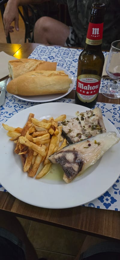 A close-up, slightly angled shot captures a meal on a table, likely at Venta de la Romera, Spain. The focus is on a white plate holding two pieces of cooked fish, possibly sea bass, seasoned with herbs and served with a side of golden-brown french fries. A white oval dish to the left contains two baguette halves. To the left of the dish and plate, a brown glass bottle of Mahou beer with a red and yellow label is prominently displayed. The label features the Mahou logo and text including "CERVEZA ESPECIAL MADRID" and "CINCO ESTRELLAS 1890". The table is covered with a white cloth adorned with a blue geometric pattern. The background shows parts of a wooden table and a person's arm and leg. The lighting suggests it could be daytime or a well-lit indoor setting. The overall impression is of a casual meal.
