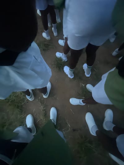 The image depicts a group of individuals, mostly seen from the knees down, wearing white outfits and white shoes. They are standing on a dirt ground with some sparse green grass. The scene appears to be outdoors, possibly in a communal or training setting. The lighting suggests it may be dusk or dawn. The individuals' attire points towards a uniform or a specific type of event. The presence of many individuals in similar clothing in an open area might indicate a gathering, ceremony, or exercise. The ground is uneven and appears to be a natural terrain.