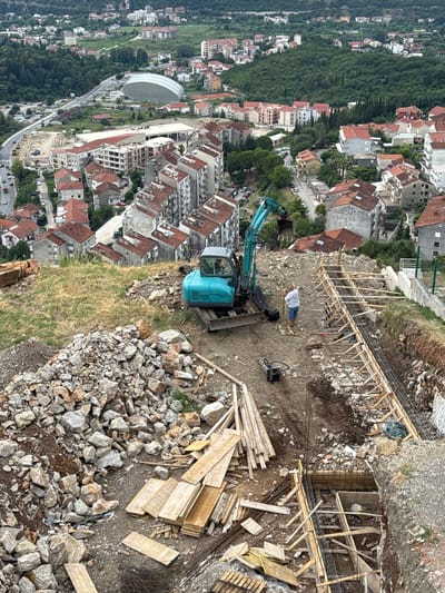 The image captures a construction site on a hillside overlooking a town with red-tiled roofs in Sušćepan, Montenegro. The scene is dominated by the construction activity, with wooden scaffolding and rebar structures in place, suggesting the early stages of building. A teal-colored excavator is positioned on the site, its arm extended, likely involved in excavation or material movement. Piles of rocks and construction debris are scattered across the ground, indicative of ongoing work. In the background, a person in a white shirt and blue jeans is lying on the ground, possibly resting or working. The surrounding landscape is characterized by lush green vegetation and the hilly terrain of Montenegro. The town below displays a dense collection of buildings, with a distinct modern, curved white structure visible in the mid-distance. The lighting suggests it is daytime, with overcast or diffused sunlight. The overall impression is one of active construction amidst a scenic, established urban environment.